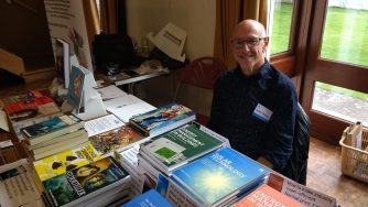 David smiling behind a stall covered with his books, including graphic novels Captain Britain and Doc Chaos, eco-thrillers Stormteller and Hybrids, and non-fiction titles Solar Technology and Energy Manangement in Buildings